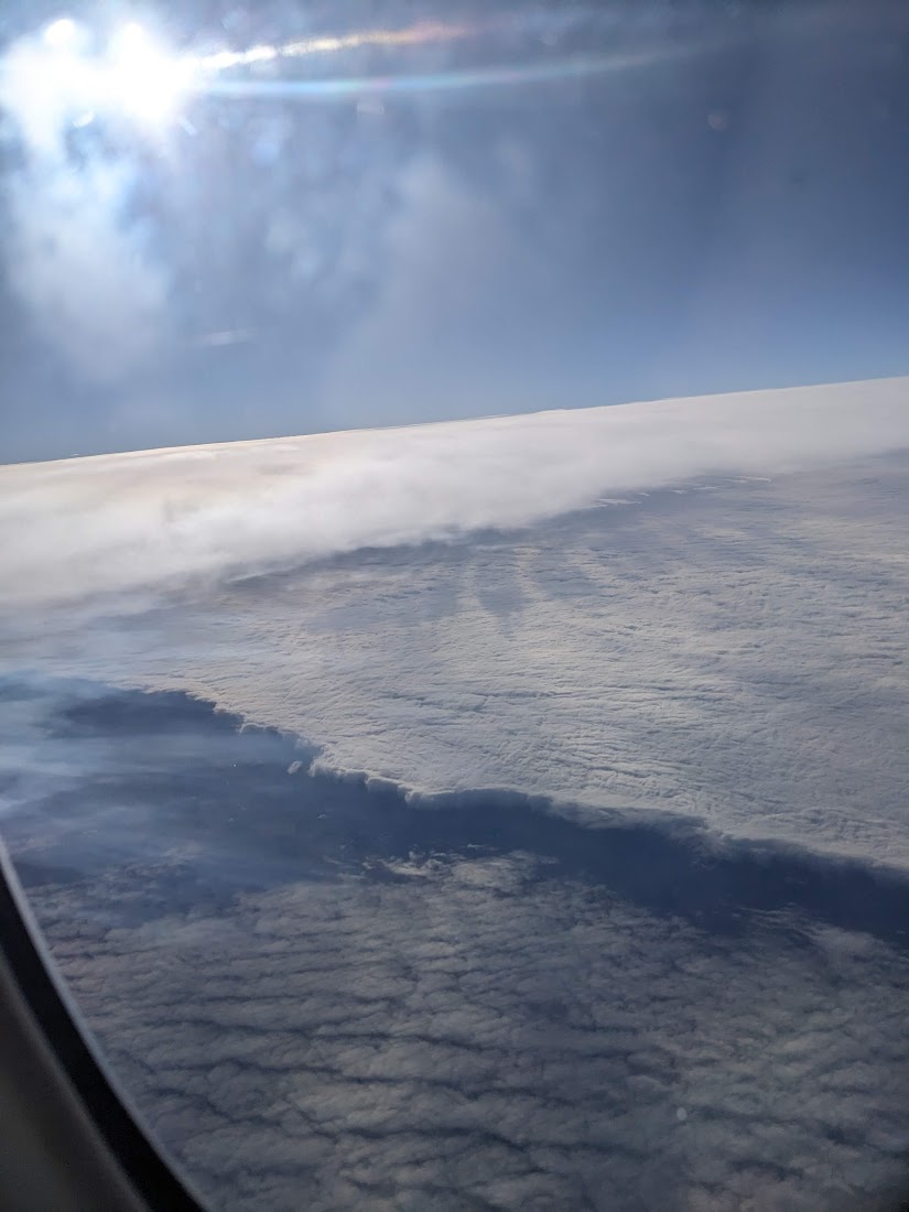 a picture taken from an airplane windows. in the top left is the sun, blurred into an incomprehensible shape by the window pane. to the right is empty space. below are layered clouds, that look almost like ocean waves.